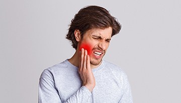 A young male wearing a gray shirt and holding his cheek that is radiating in pain because of a severe toothache