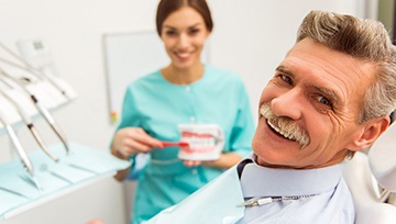 An older man smiling while a dental hygienist shows him how to care for his dentures in the background