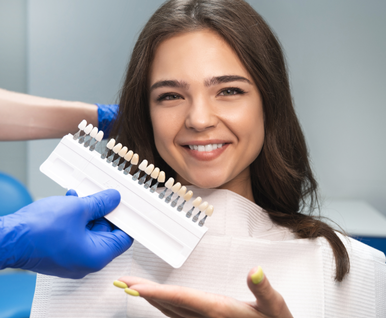 Woman in dental chair smiling with hand showing set of veneer samples